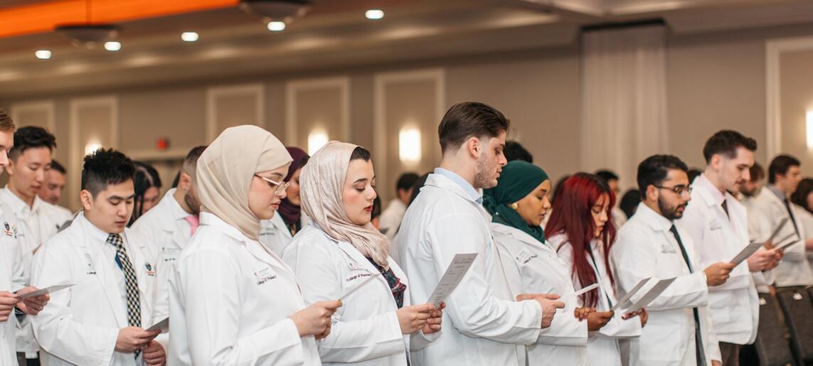 A lineup of students read the pharmacists oath at a white coat ceremony, all wearing white coats, from various backgrounds.