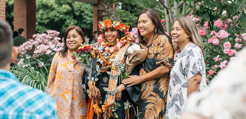A family and their graduate smile as they have their photo taken by someone aside. The graduate wears a series of leis, flowers, and more over her robes.