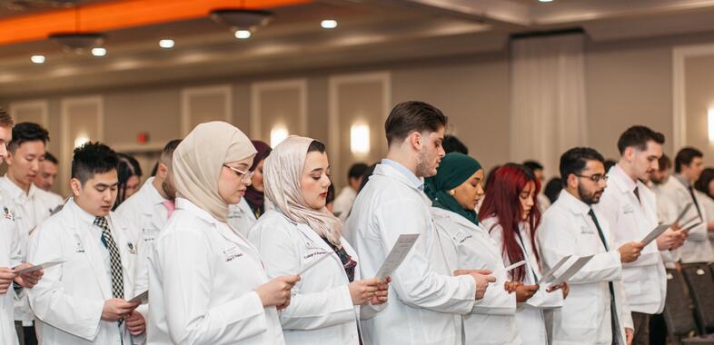 A lineup of students read the pharmacists oath at a white coat ceremony, all wearing white coats, from various backgrounds.