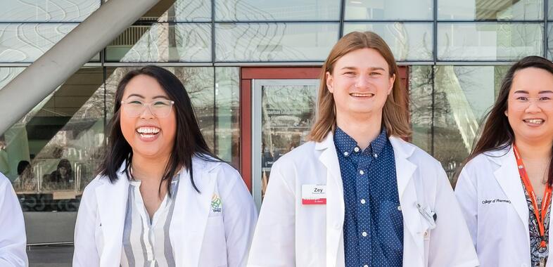 four pharmacy students wearing white coats smile while walking outside towards the camera