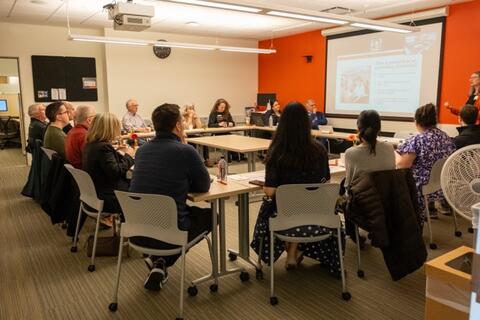 A group of alumni sit around a series of tables in a circle while watching a presentation